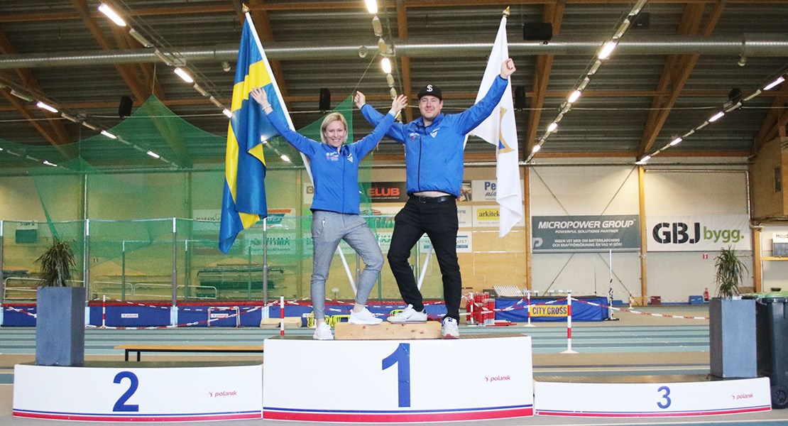 Micaela Wieslander Meyer och David Stridfeldt står överst på en prispall i Telekonsult Arena och sträcker händerna i luften i en segergest.