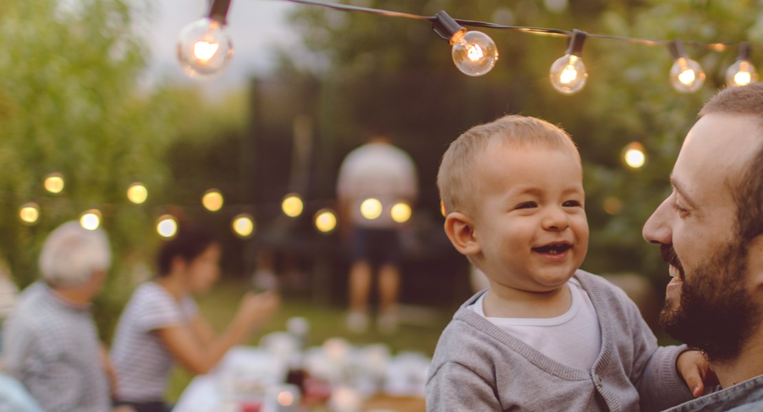 Man som står under en ljusslinga utomhus och håller ett skrattande barn i famnen, i bakgrunden syns människor runt ett bord i en trädgård.