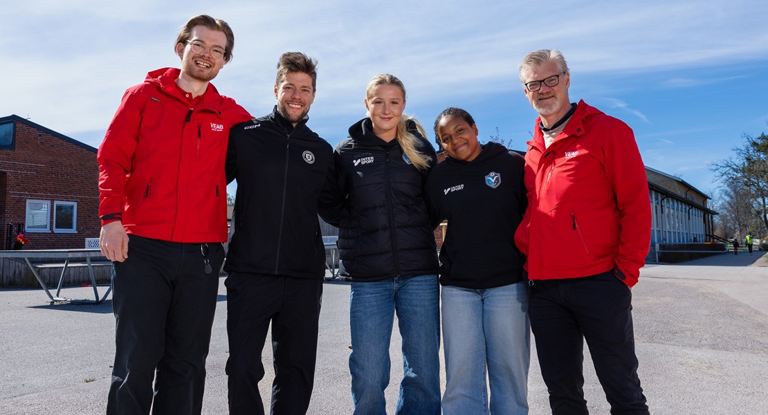 Fem personer står på rad och ler mot kameran, vid en skolgård med blå himmel.