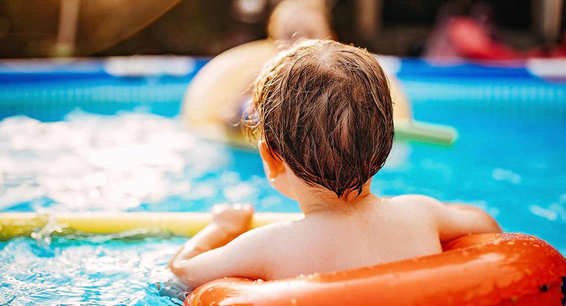 Glad pojke i blå badbyxor och med simglasögon i pannan ligger i en rosa badring i en pool.