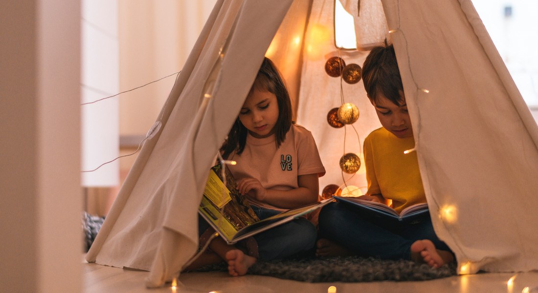 Två barn sitter i ett tält inomhus och läser bok